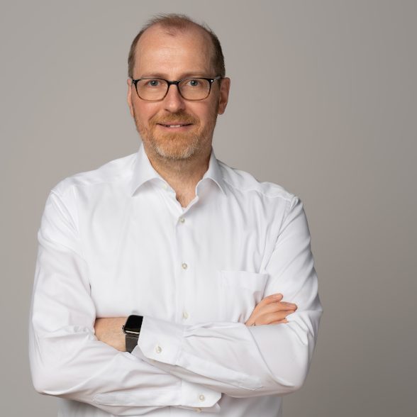 Mann mit Brille trägt ein weißes Hemd, steht mit verschränkten Armen vor neutralem Hintergrund.
